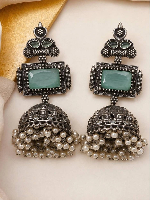 Front-facing pair of oxidised silver jhumka earrings with faceted green stone and pearl dangles on cream background
