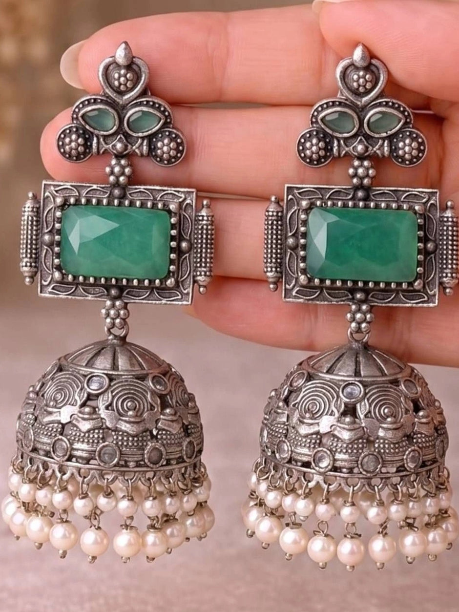 Close-up of oxidised silver jhumka earrings with green stone held in hand showing detailed temple engravings and pearl fringe