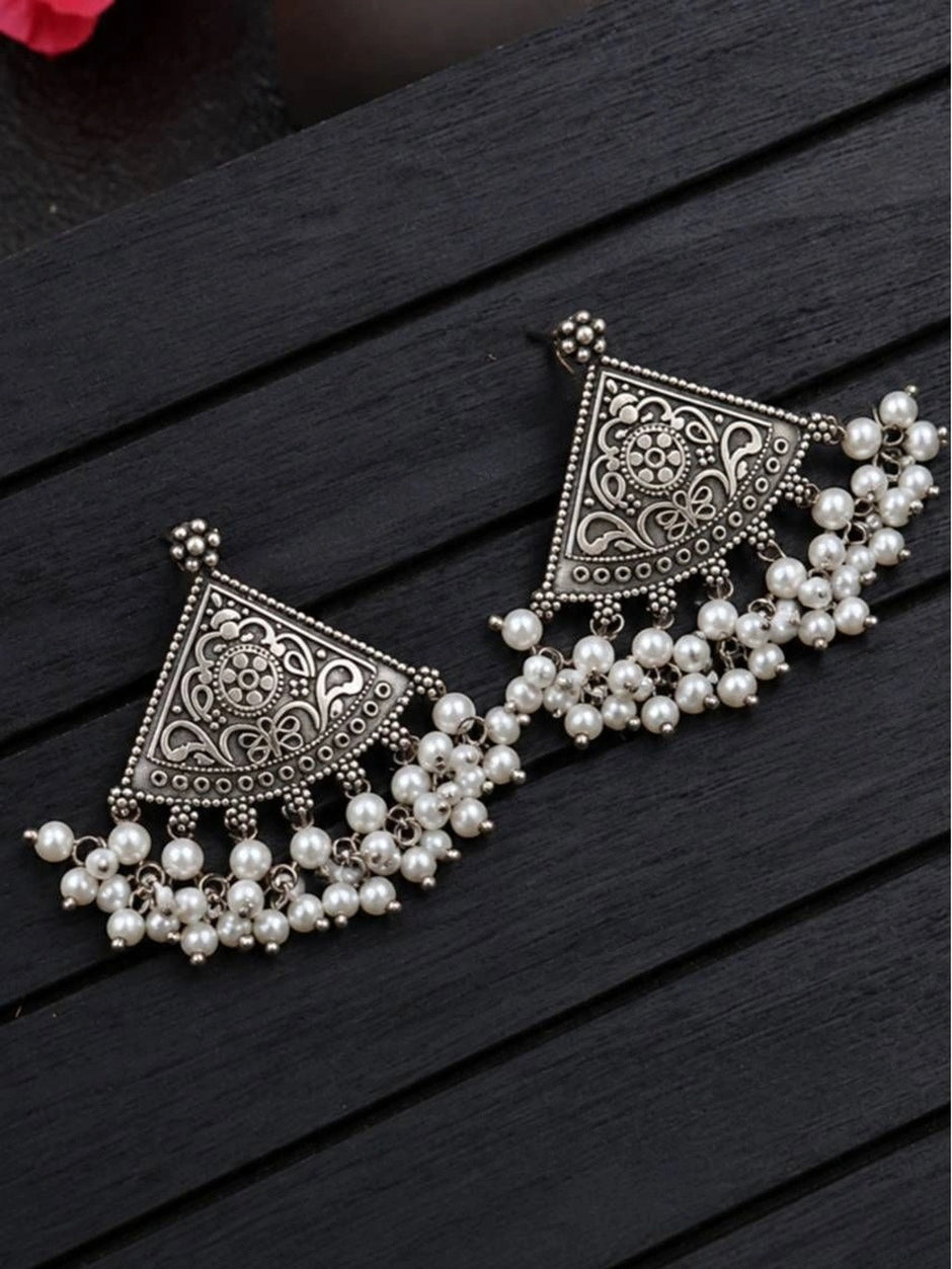 Two oxidised silver chandbali earrings with engraved floral motifs and pearl clusters laid flat on dark wood