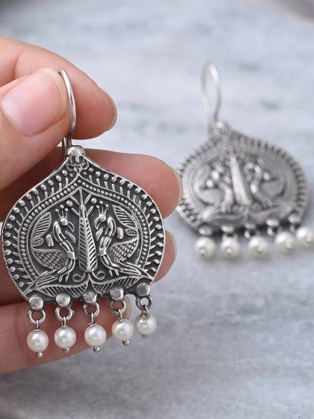 Close-up of oxidised silver peacock earring held between fingers showing detailed engraving and pearl drops