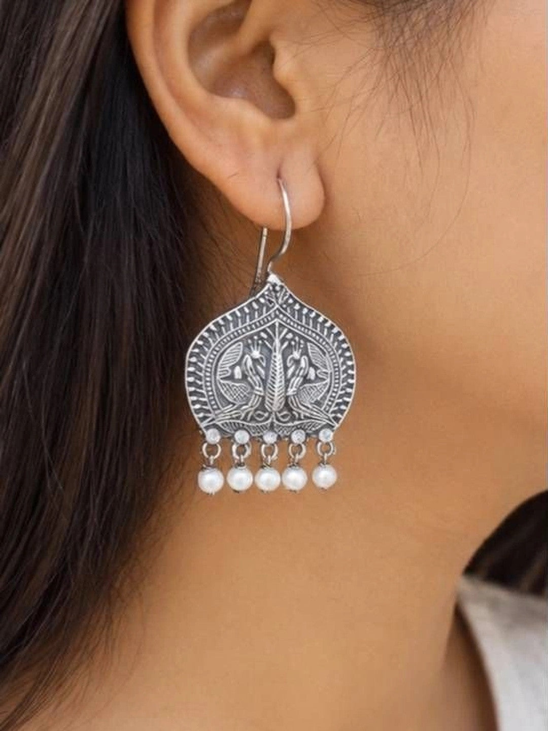 Oxidised silver peacock drop earring with white pearls worn on a woman's ear against light background