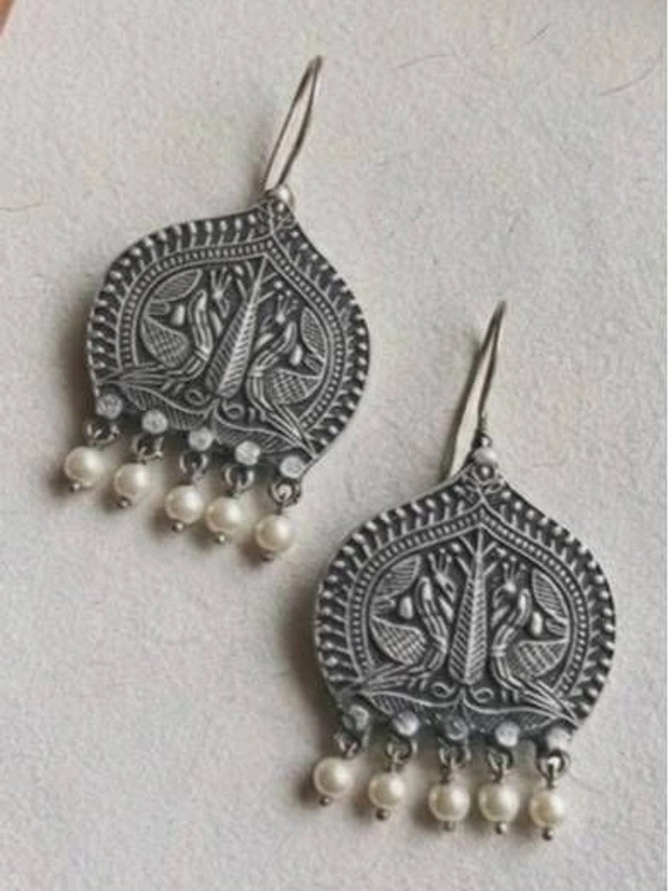 Two oxidised silver peacock earrings with pearl drops laid flat on a white textured surface