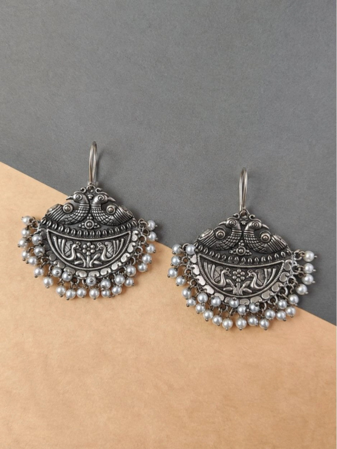 Pair of oxidised silver chandbali earrings with peacock crest and white pearl bead fringe on neutral background