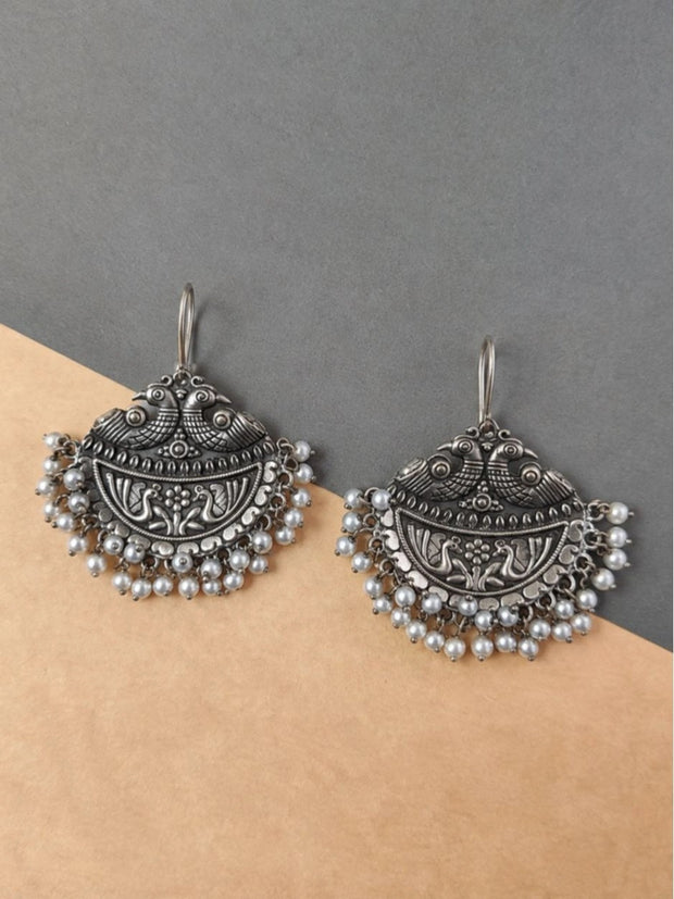 Pair of oxidised silver chandbali earrings with peacock crest and white pearl bead fringe on neutral background