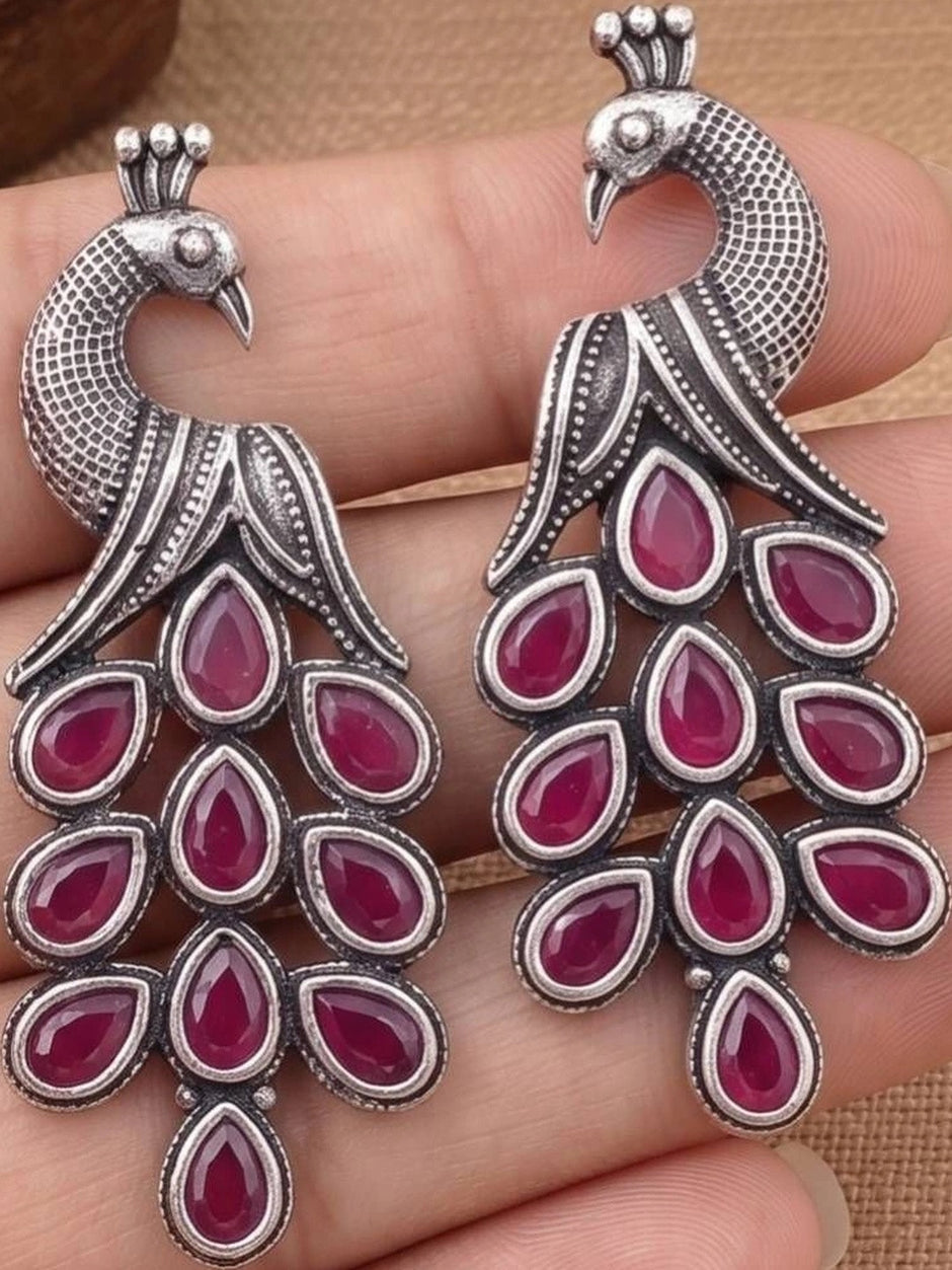 Pair of oxidised silver peacock drop earrings with ruby-red teardrop stones held in hand