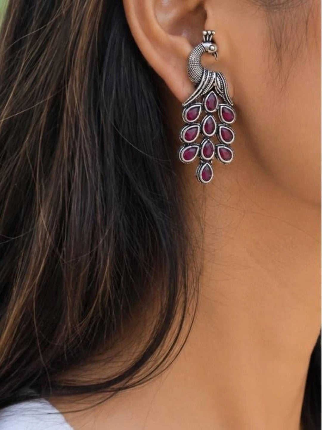 Oxidised silver peacock drop earring worn on woman's ear against dark hair