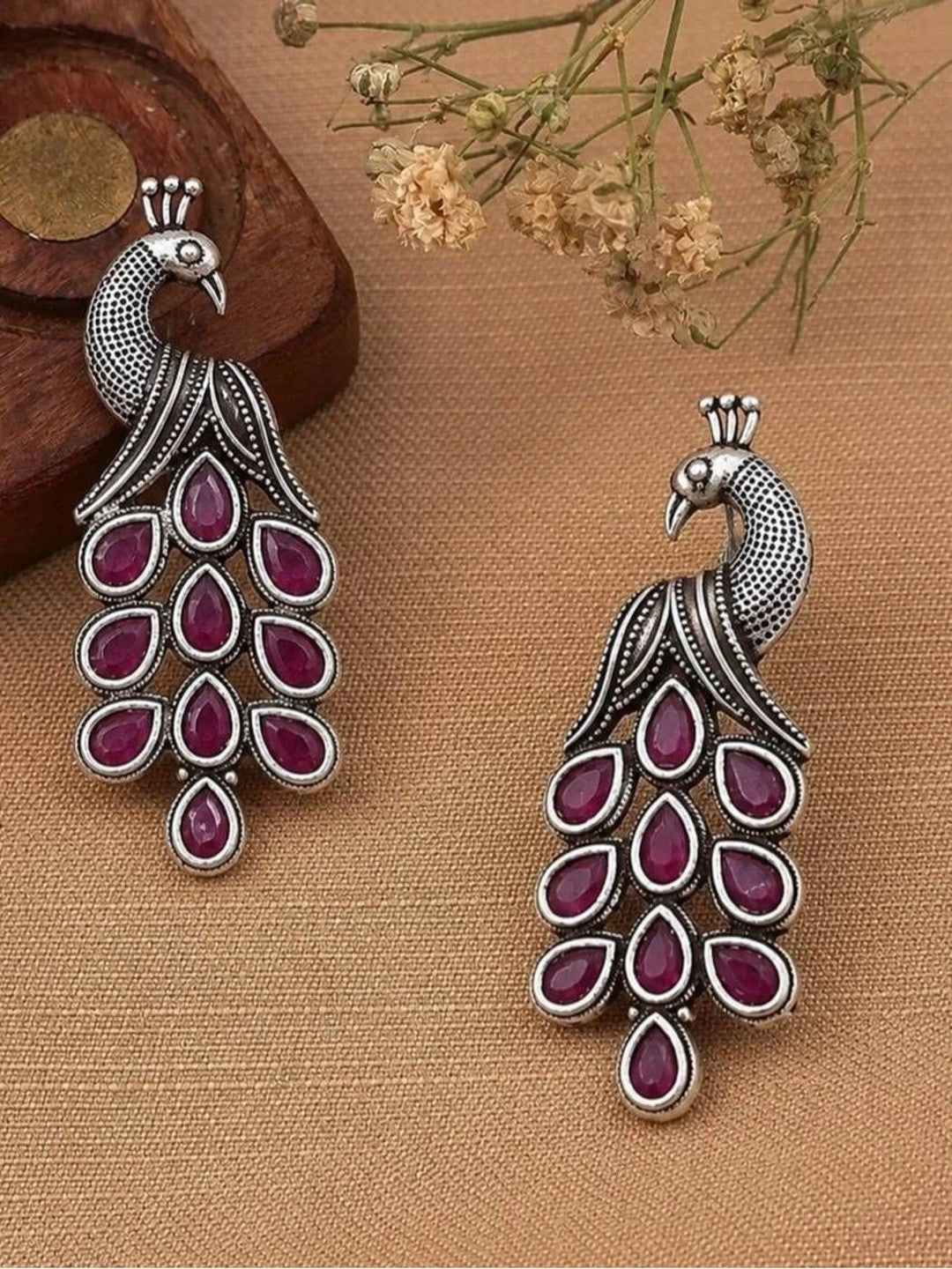 Pair of oxidised silver peacock earrings with ruby-red stones displayed on beige fabric