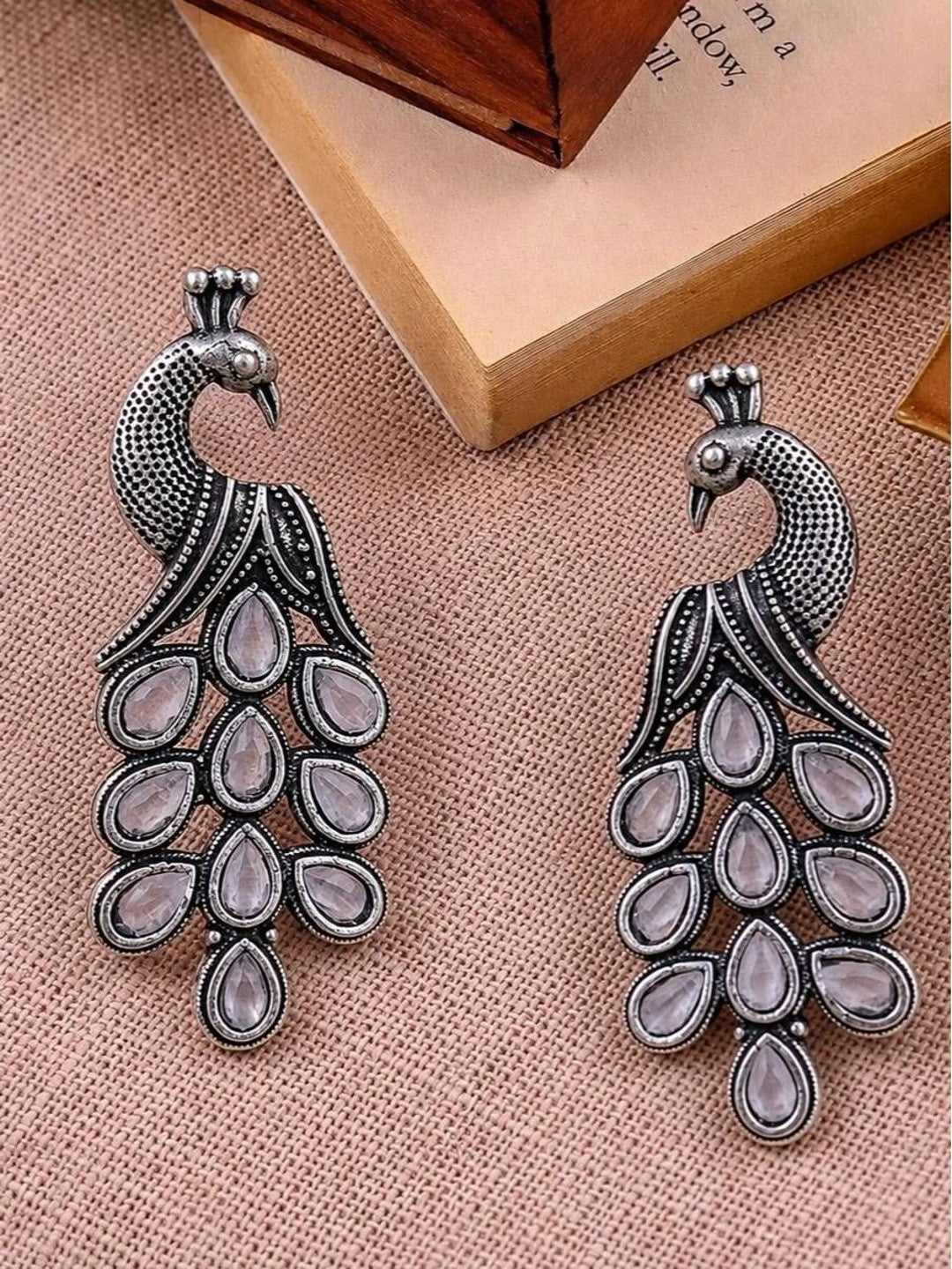 Pair of oxidised silver peacock drop earrings laid flat showing mirrored teardrop tail stones
