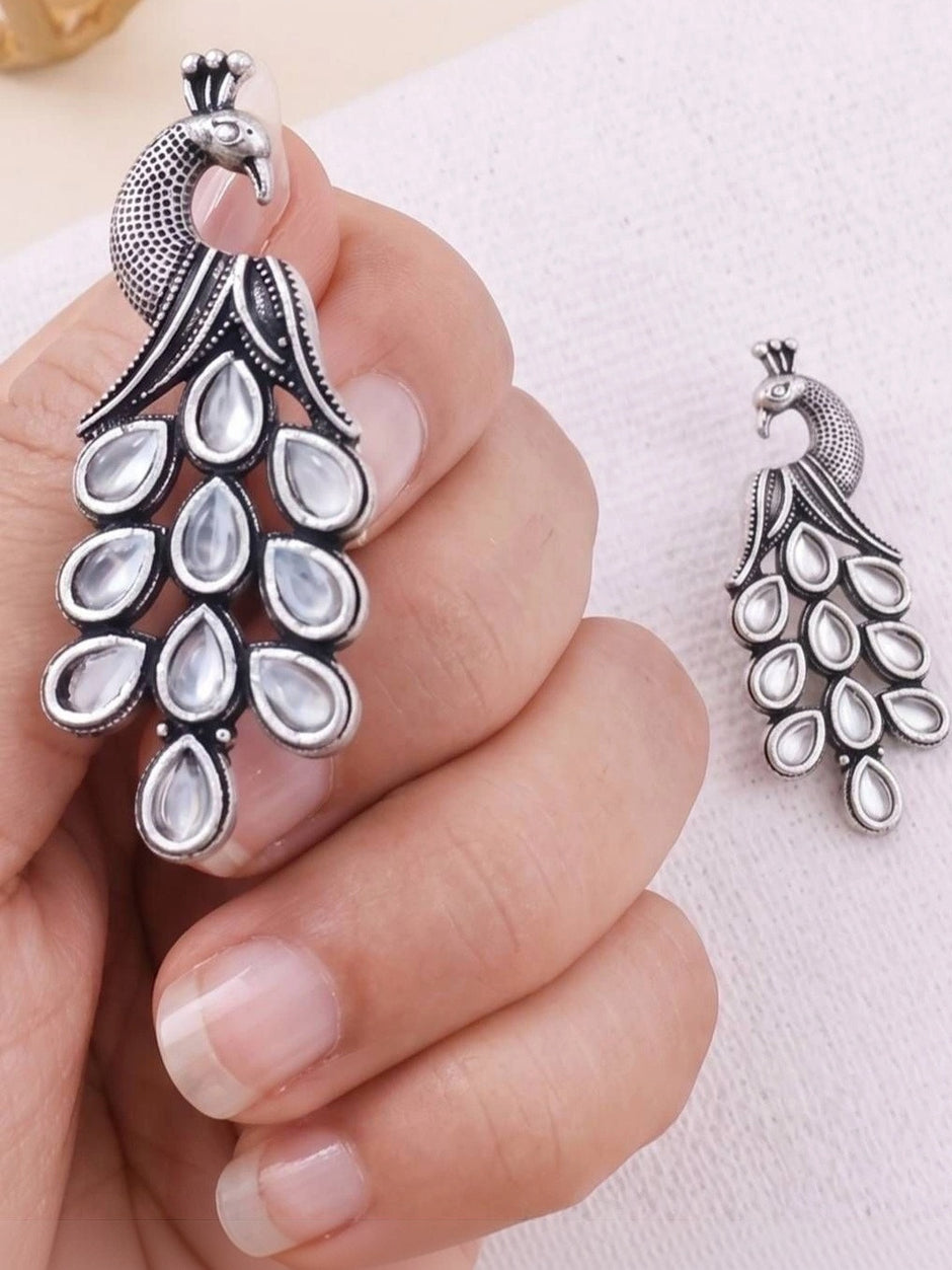Hand holding a large oxidised silver peacock earring with clear kundan teardrop stones alongside the smaller matching earring