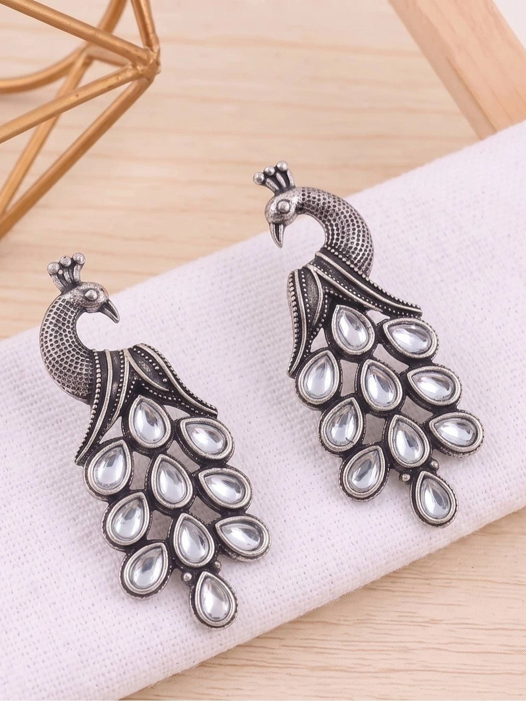 Pair of oxidised silver kundan peacock earrings laid flat on white fabric with gold geometric decor in background