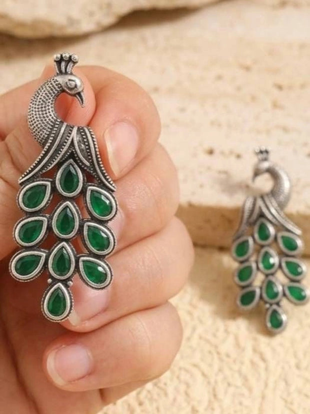 Hand holding a large oxidised silver peacock earring with green teardrop stones cascading as the tail