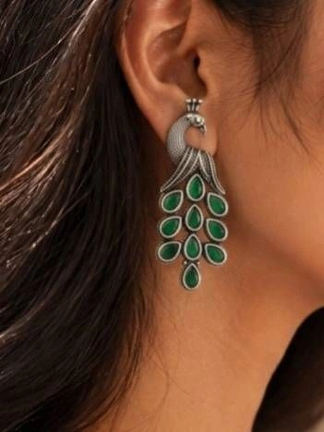 Oxidised silver peacock drop earring worn on a woman's ear showing full length and green stone tail