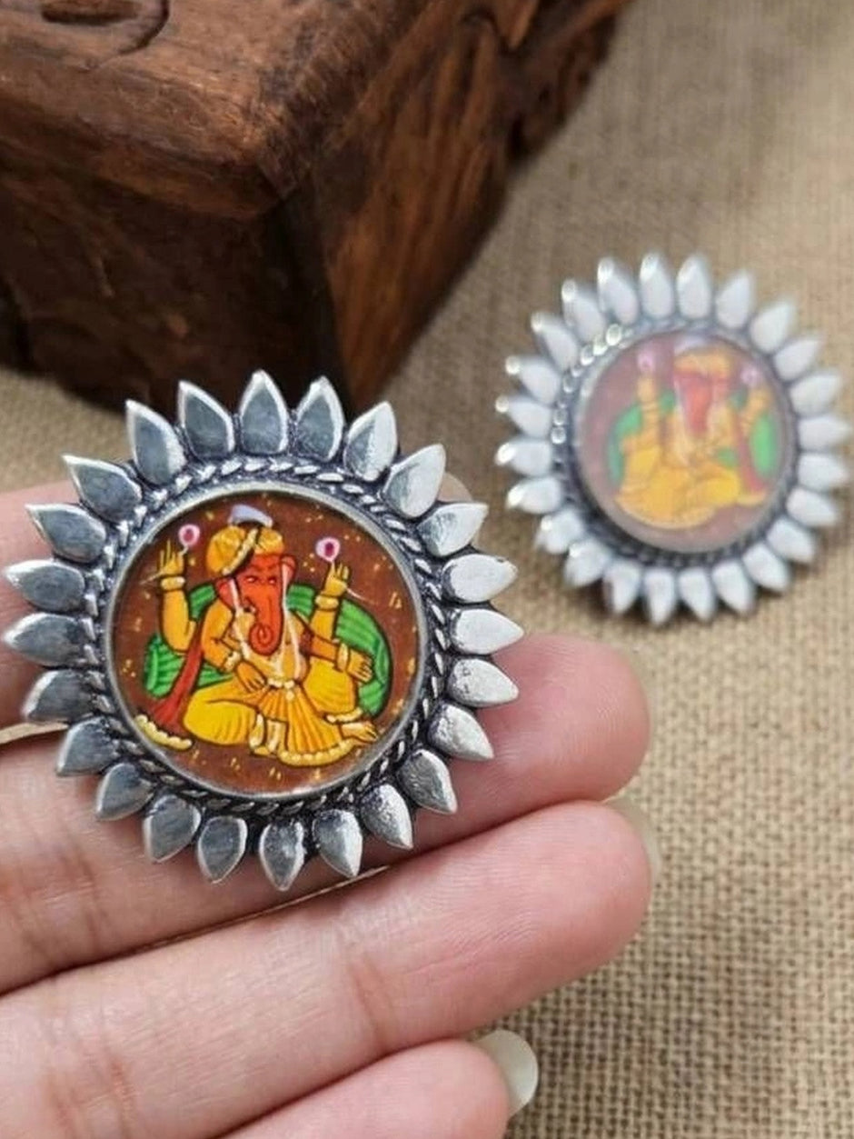 Pair of oxidised silver Ganesha stud earrings held in a woman's palm showing sunflower-petal border and painted Ganesha motif