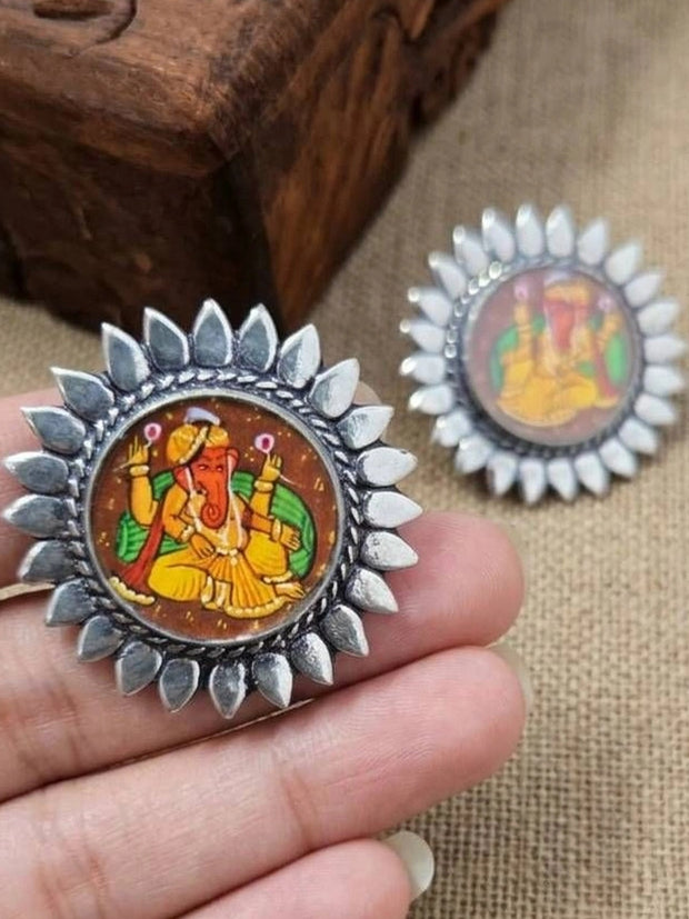 Pair of oxidised silver Ganesha stud earrings held in a woman's palm showing sunflower-petal border and painted Ganesha motif