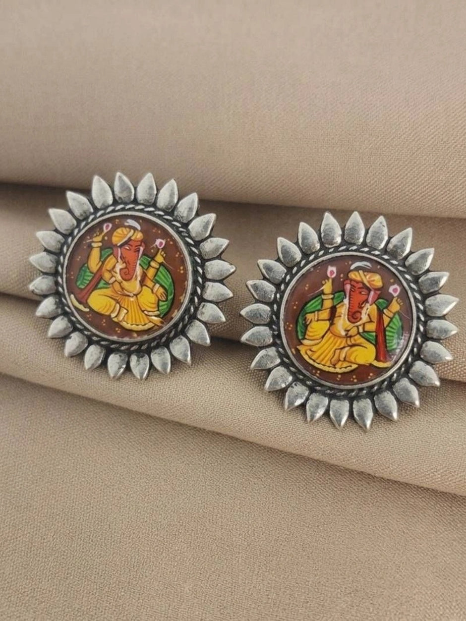 Pair of oxidised silver Ganesha stud earrings laid flat on a beige fabric showing mirrored Ganesha illustrations