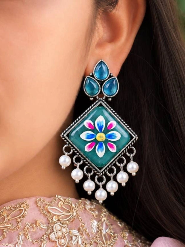 Oxidised silver Meenakari square earring worn on ear, showing teal enamel flower and pearl drops against pink embroidered outfit