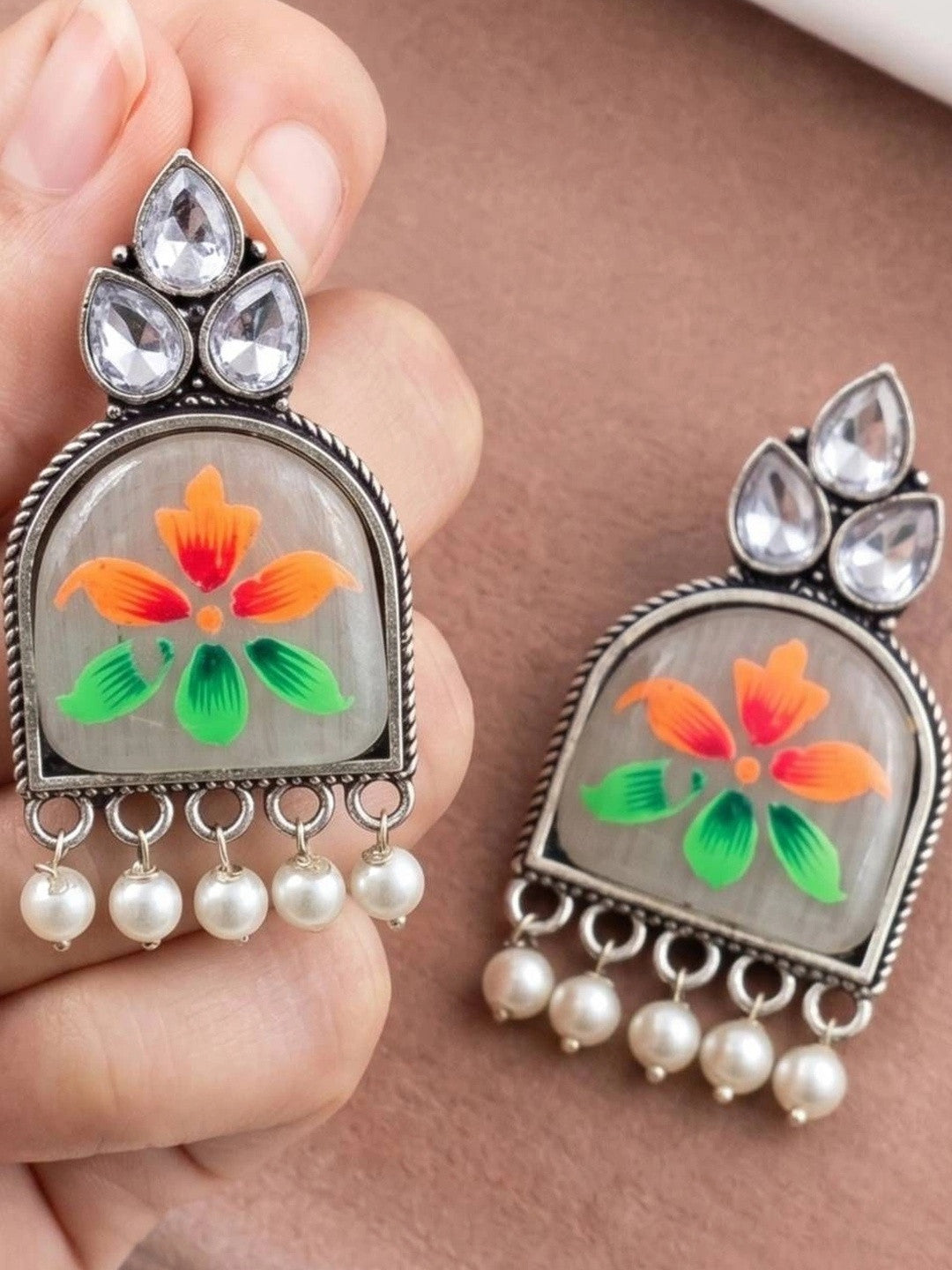 Hand holding a pair of oxidised silver meenakari chandbali earrings showing orange and green floral motif with pearl drops