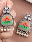 Hand holding a pair of oxidised silver meenakari chandbali earrings showing orange and green floral motif with pearl drops