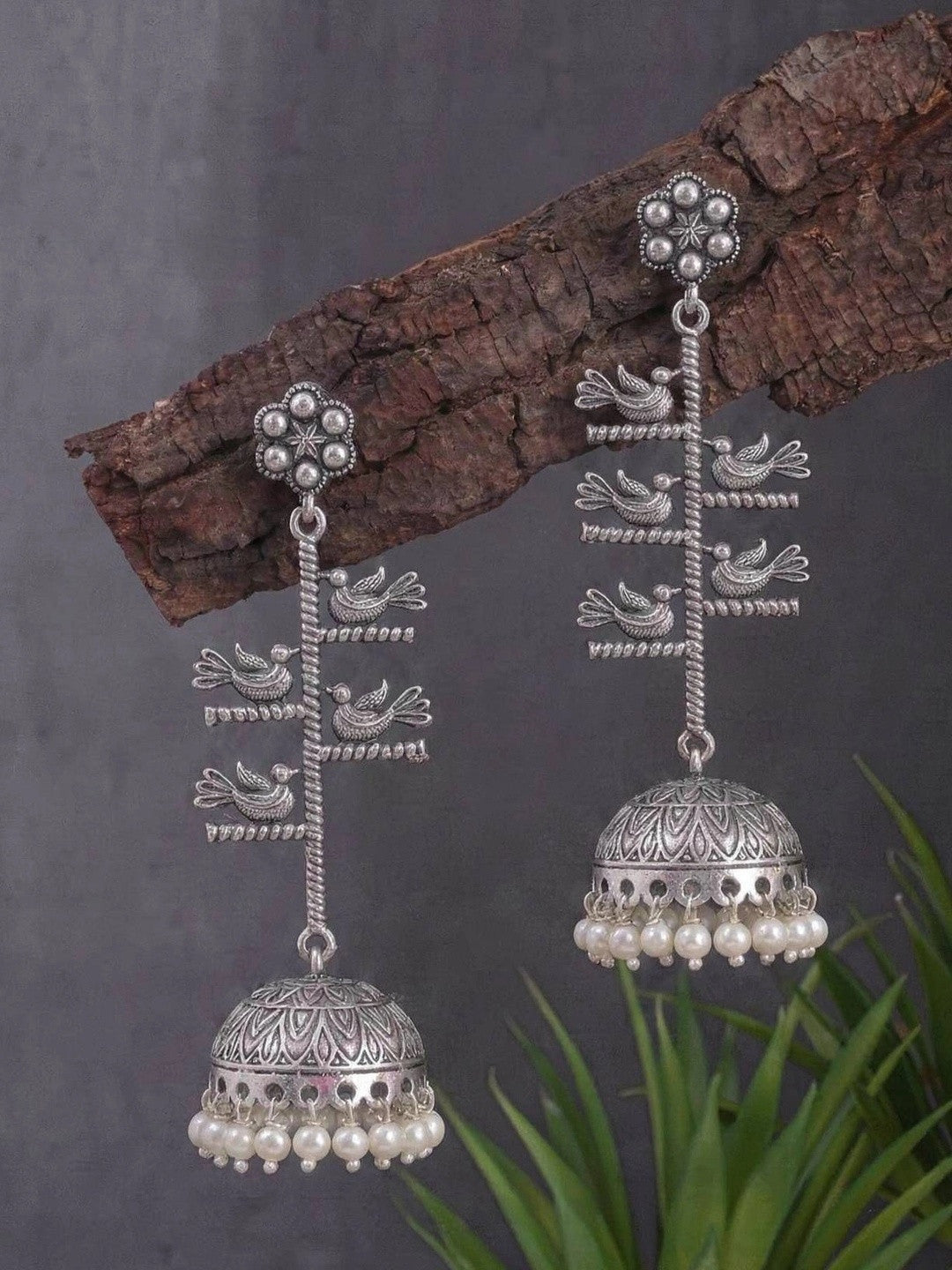 Two oxidised silver bird jhumka earrings displayed on a wooden branch against grey background