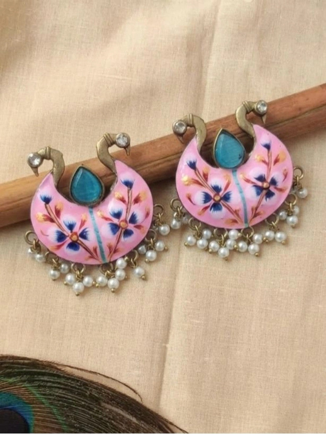 Pair of pink meenakari chandbali earrings displayed on wooden stick with peacock feather