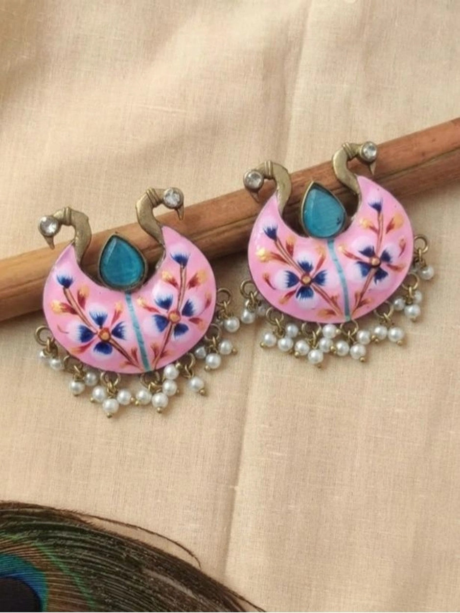 Pair of pink meenakari chandbali earrings displayed on wooden stick with peacock feather