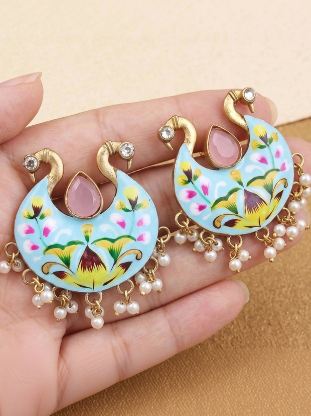 Pair of sky-blue meenakari peacock chandbali earrings with pearl drops held on a palm