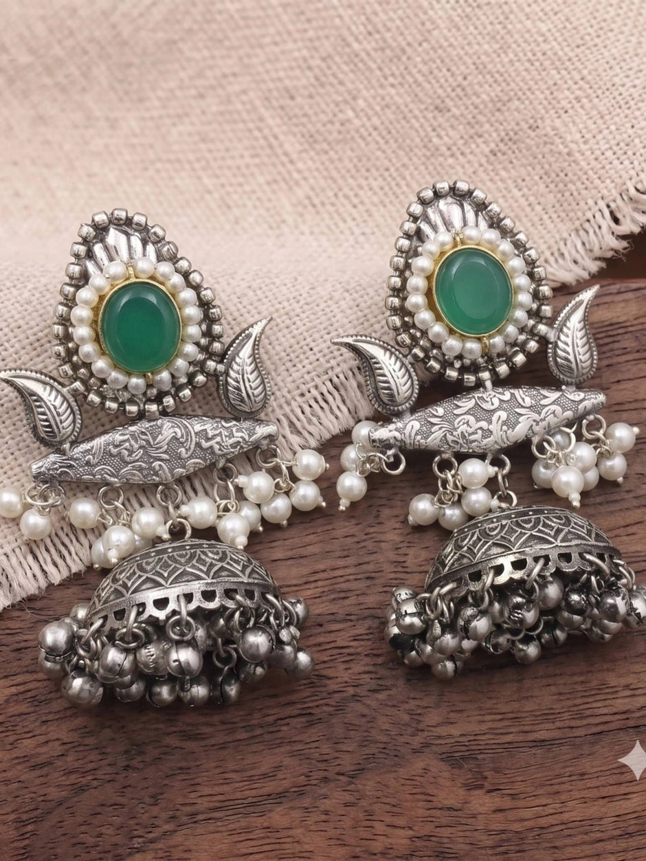 Pair of oxidised silver jhumka earrings laid flat on wooden surface showing engraved bridge, faux pearl clusters and domed bell with ghungroo