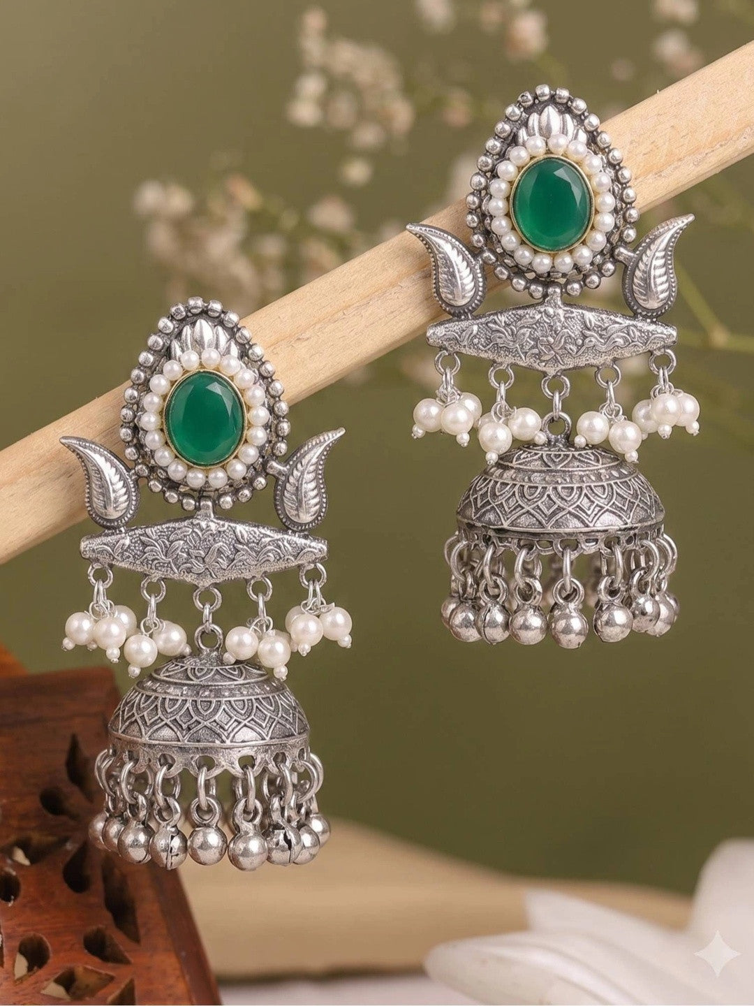 Two oxidised silver teardrop jhumkas balanced on a wooden rod against a sage green background with floral bokeh