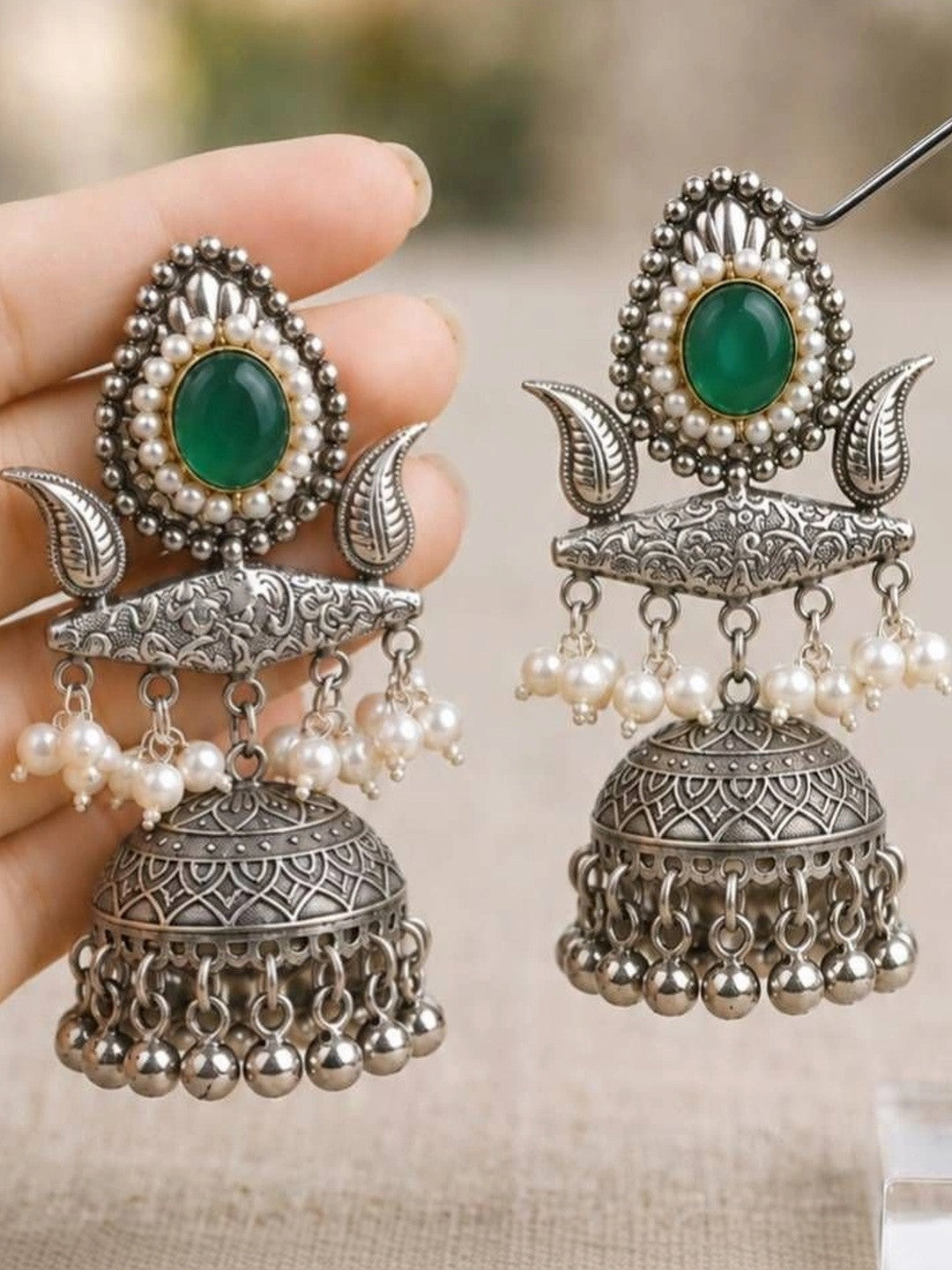 Close-up of oxidised silver jhumka pair held in hand and displayed on stand showing intricate mandala engraving and multi-tier pearl and bell drops