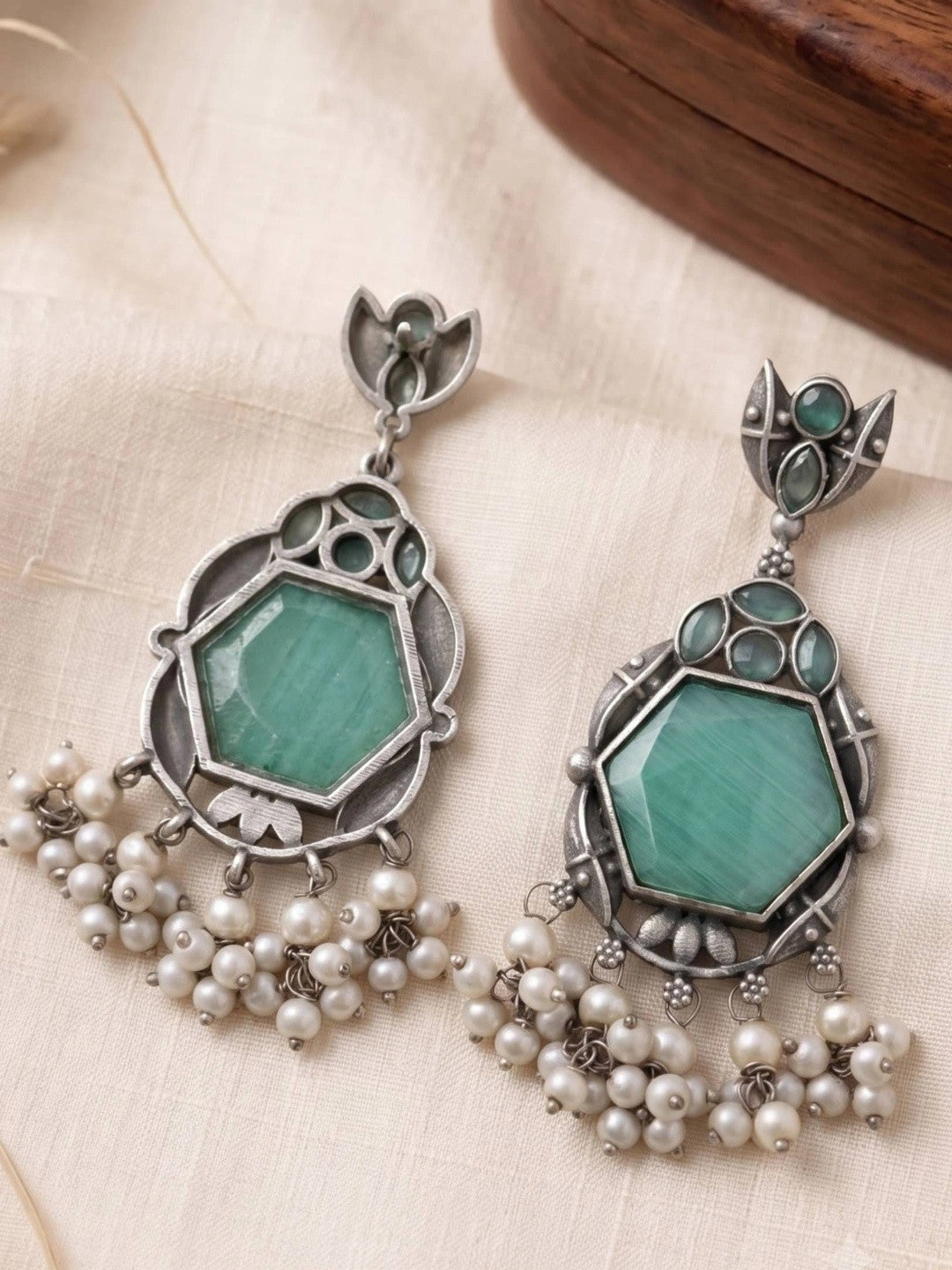 Pair of oxidised silver green stone hexagon earrings with pearl drops laid flat on cream fabric