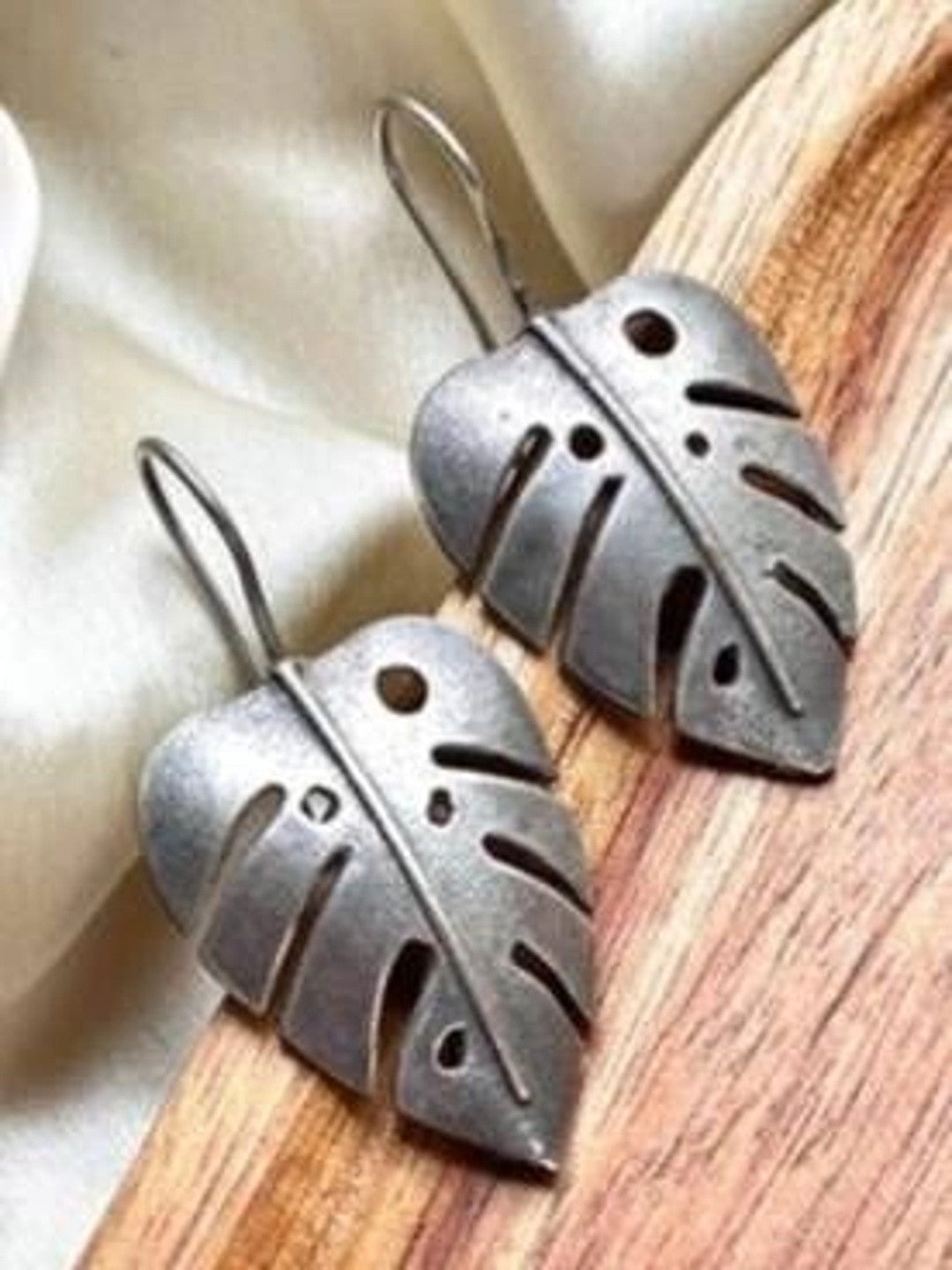 Pair of oxidised silver monstera leaf drop earrings lying flat on a wooden surface beside cream fabric