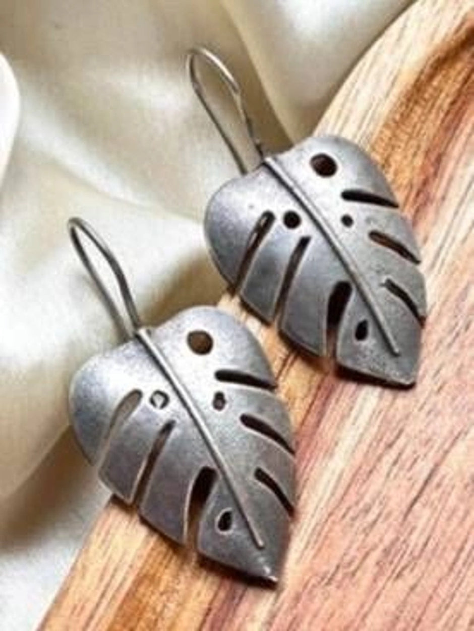 Pair of oxidised silver monstera leaf drop earrings lying flat on a wooden surface beside cream fabric