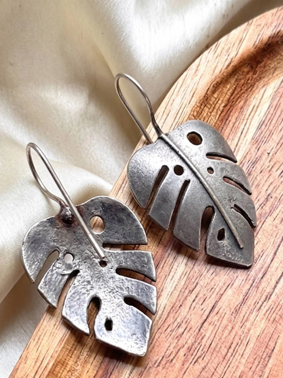 Close-up of two oxidised silver leaf earrings on a round wooden tray showing textured cut-out detail