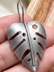 Single oxidised silver monstera leaf earring held between fingers showing full cut-out leaf shape and hook