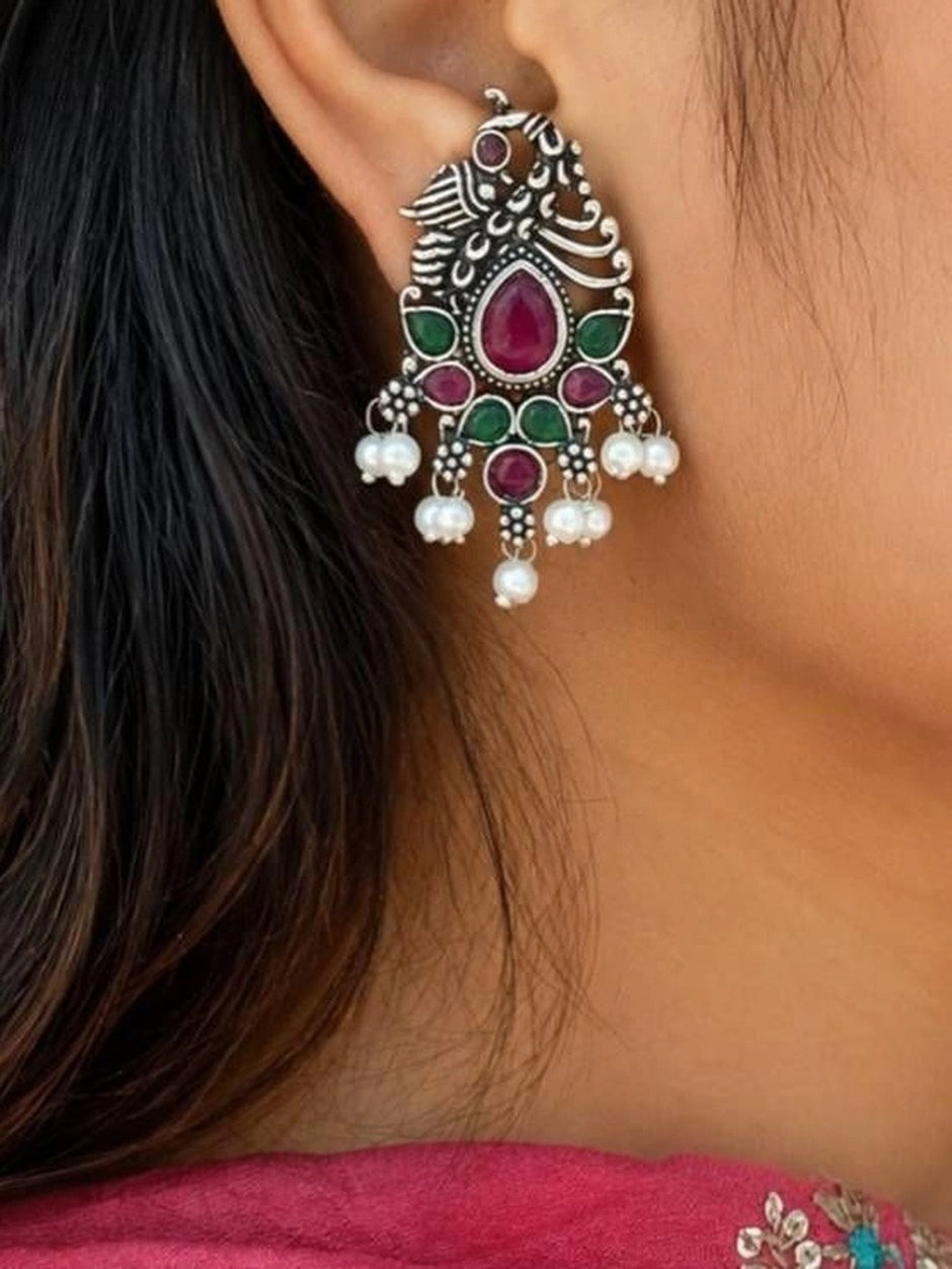 Oxidised silver peacock drop earring worn on a woman's ear with ruby and green stones