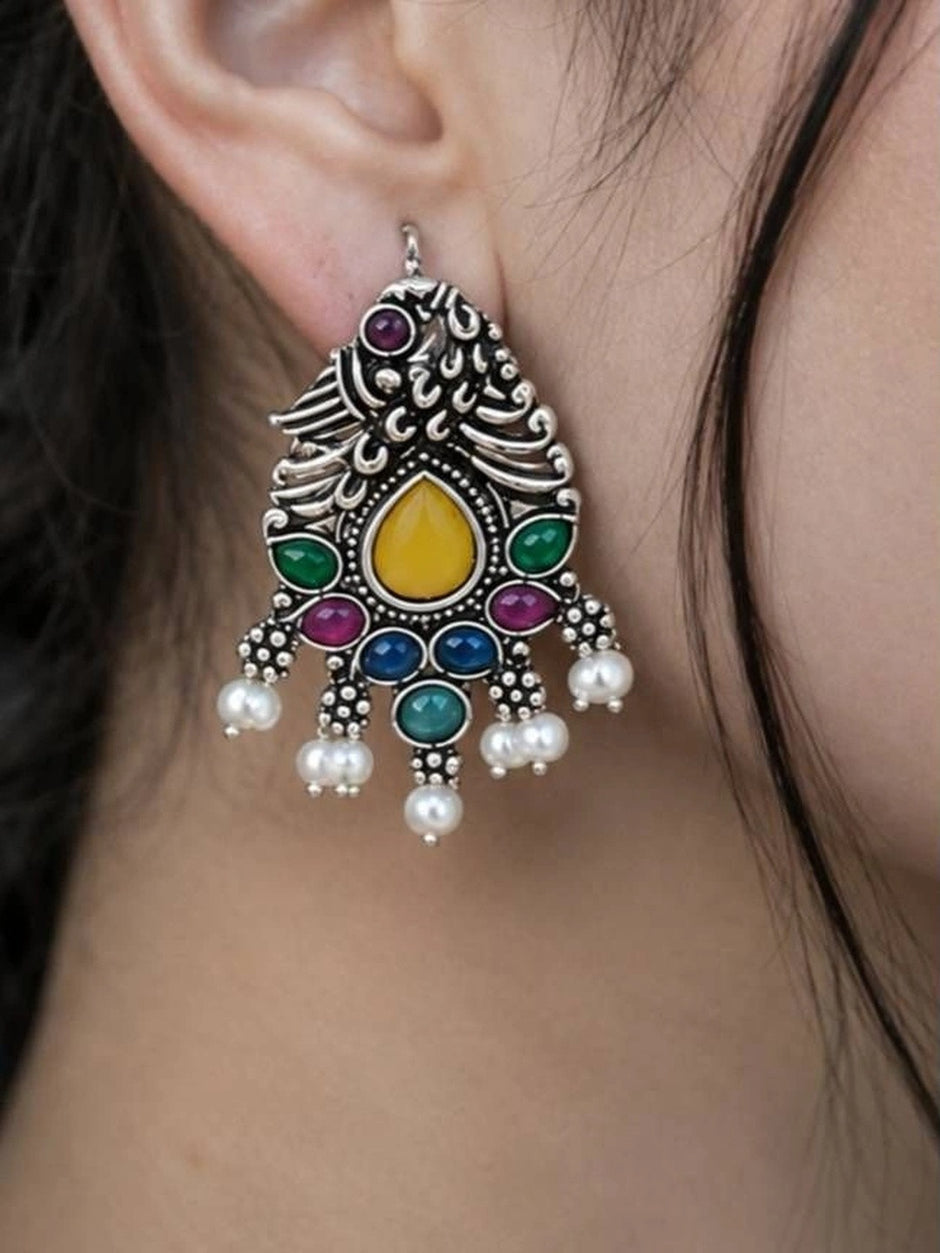 Oxidised silver peacock drop earring worn on a woman's ear, showing multicolour stones and white pearl drops