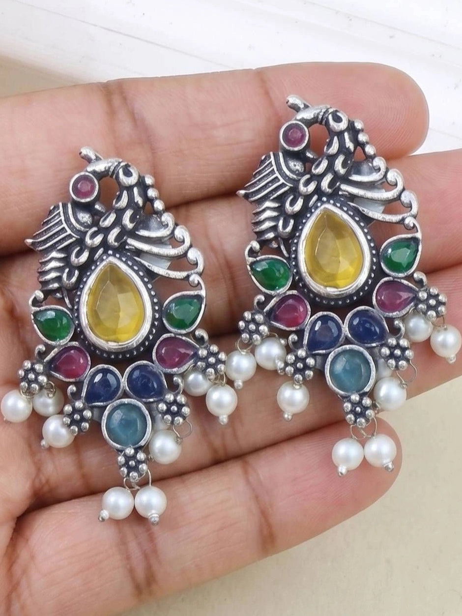 Close-up of two peacock earrings held in a hand showing coloured glass stones and pearl drops in detail