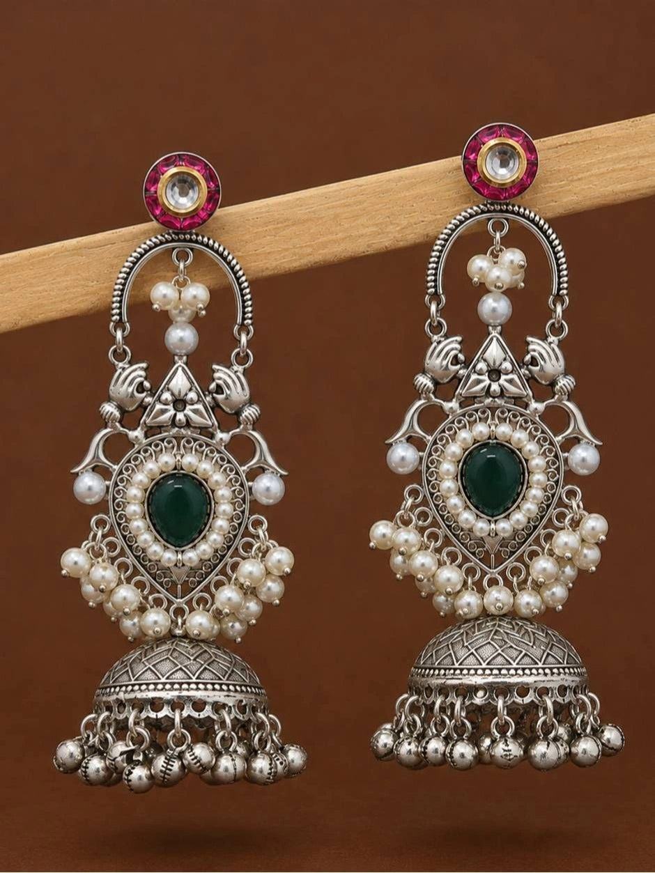 Side-by-side pair of oxidised silver chandbali jhumka earrings showing peacock motif, green stone, pearl clusters and ghungroo drops against brown background