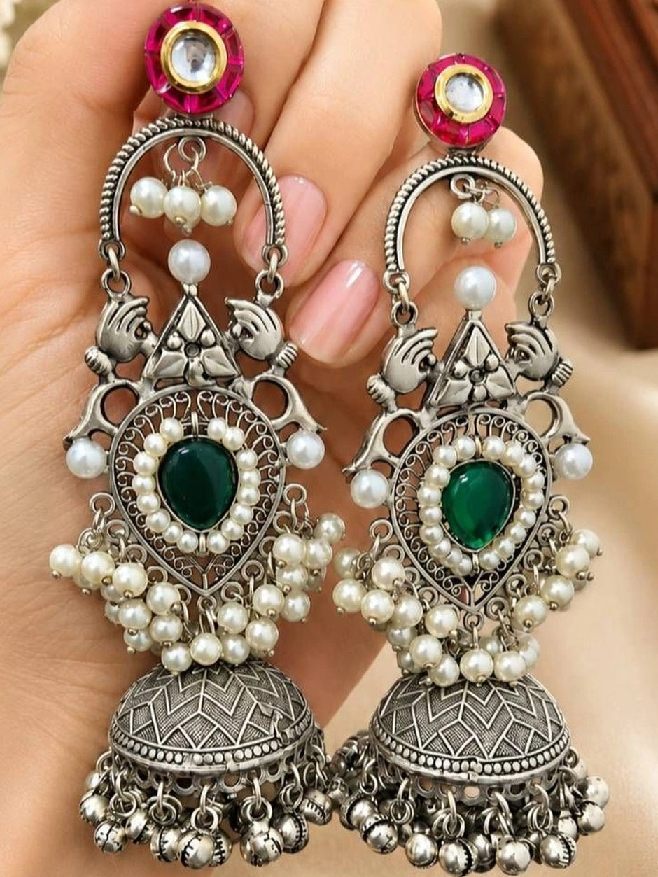 Pair of oxidised silver pearl and green stone jhumka earrings held in a woman's palm showing filigree detail and peacock motif up close
