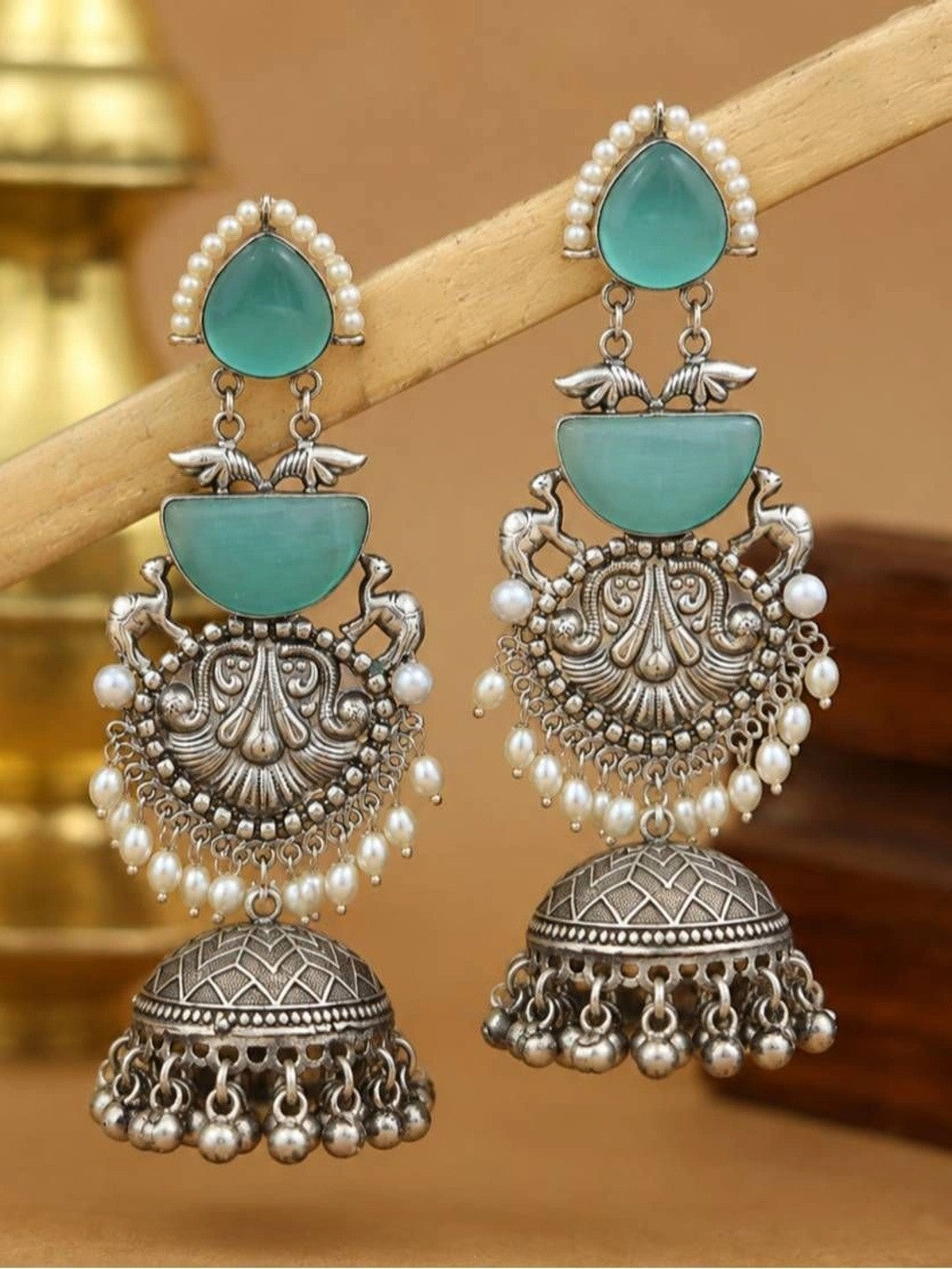 Pair of layered turquoise and oxidised silver jhumka earrings resting on wooden prop with brass vessel background