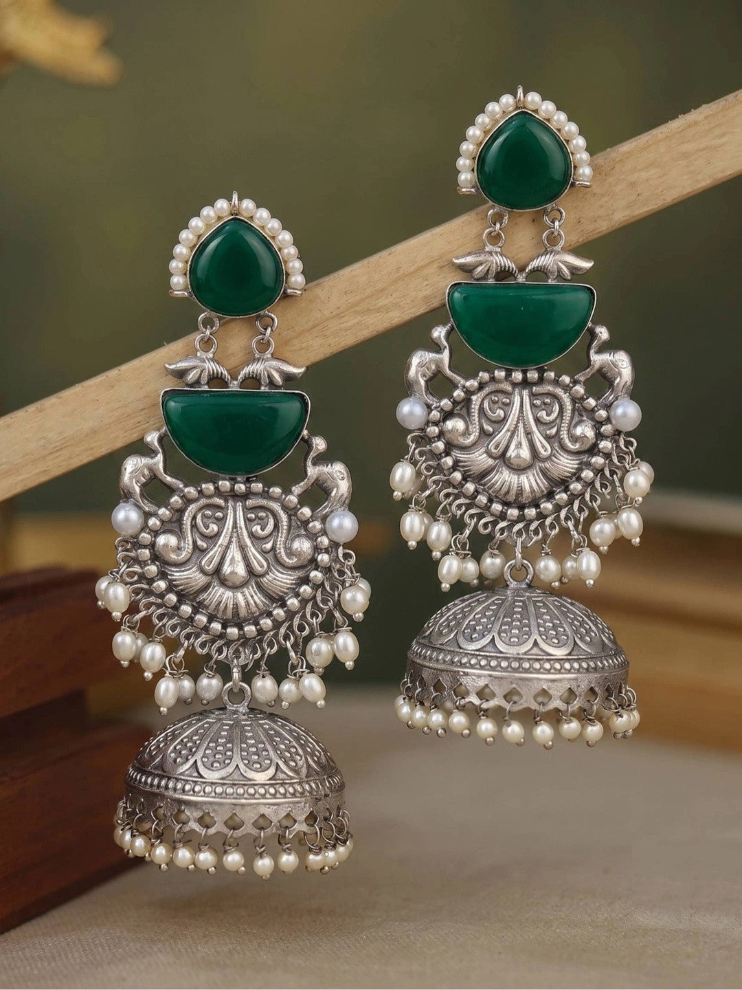 Two oxidised silver green stone jhumkas with peacock motifs resting on a wooden stand