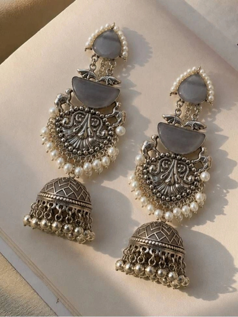 Two oxidised silver layered jhumka earrings laid flat on a cream surface showing full design detail