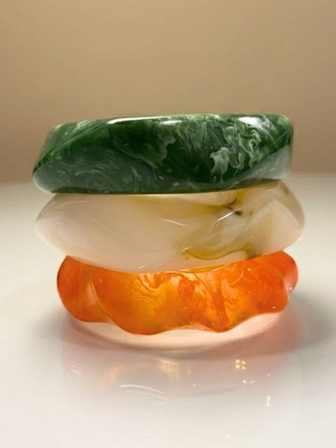 Three stacked resin bangles in green marble, pearl white, and orange on a white surface