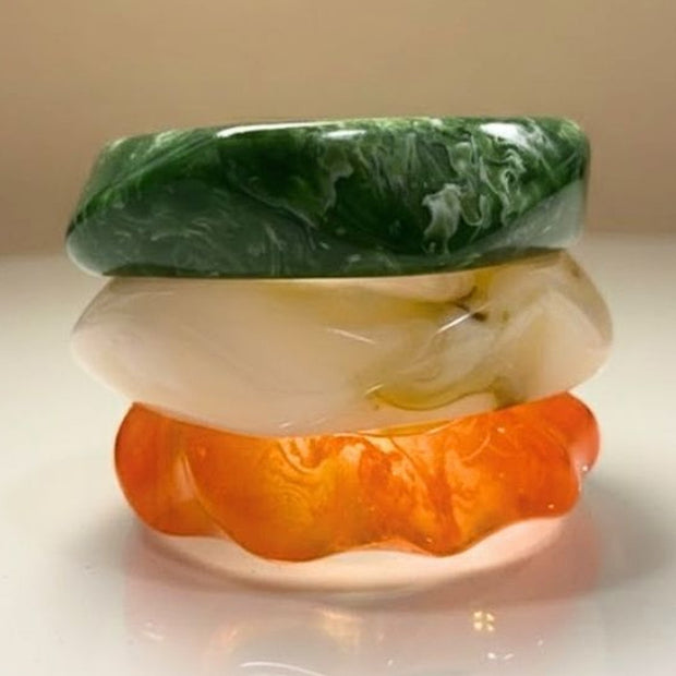 Three stacked resin bangles in green marble, pearl white, and orange on a white surface