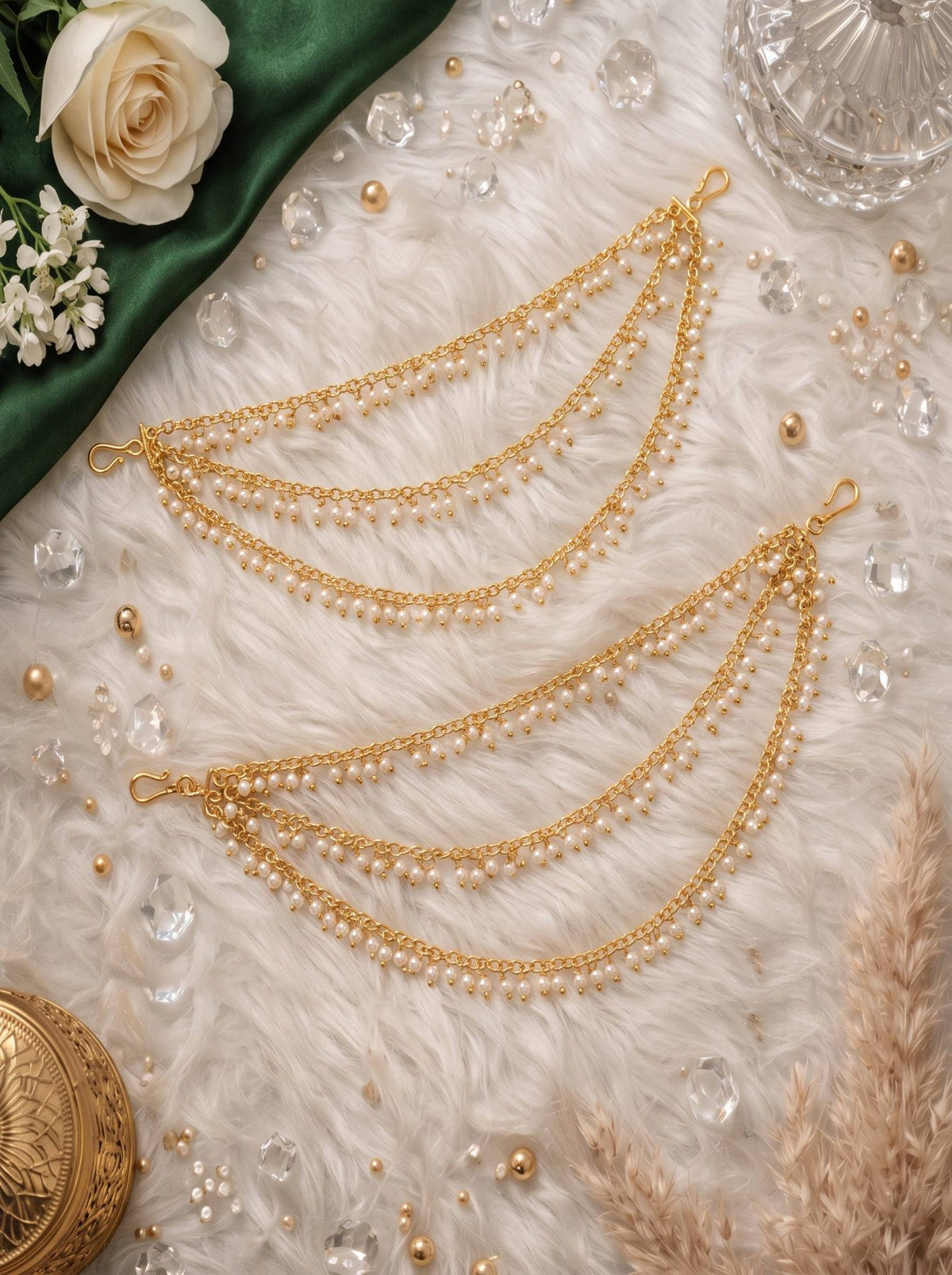 Pair of gold-plated three-layer pearl ear chains laid flat on white fur with crystals and a white rose
