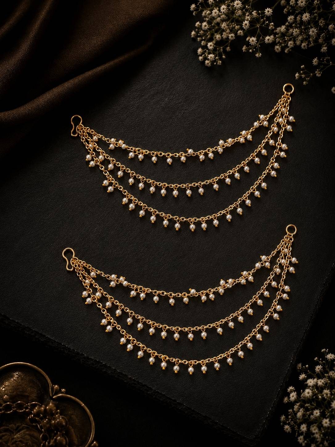Two gold-plated layered pearl ear chains displayed on a dark black surface with baby's breath flowers