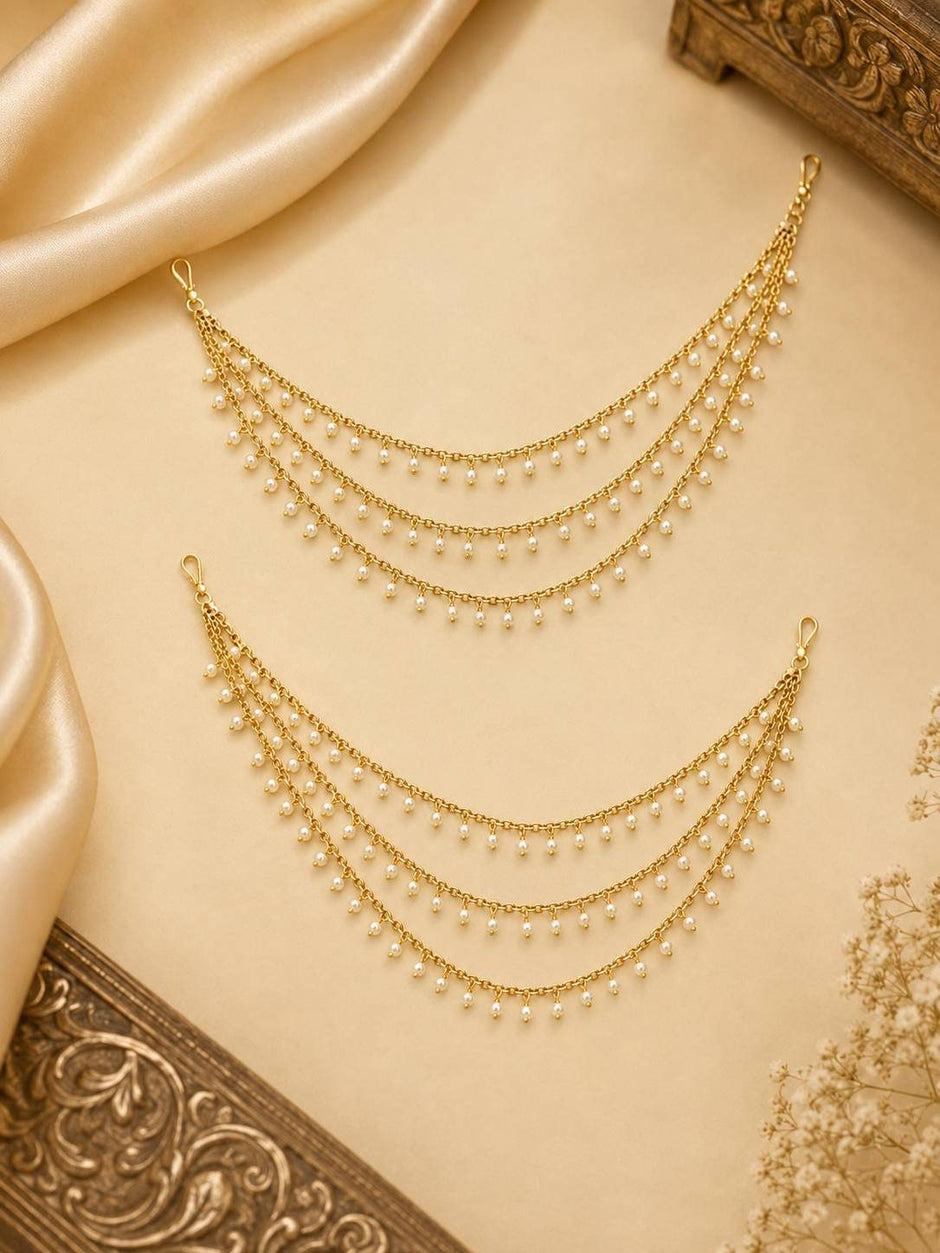 Pair of gold-plated pearl drop ear chains laid on cream satin fabric beside ornate brass decor