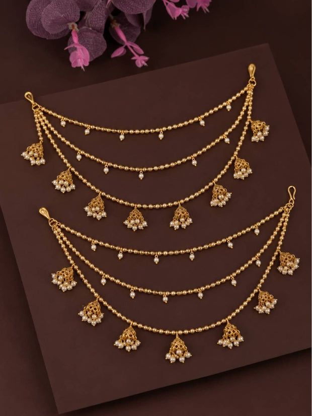 Two pairs of gold-plated multi-strand jhumka ear chains with pearl drops laid flat on dark brown surface with pink flowers