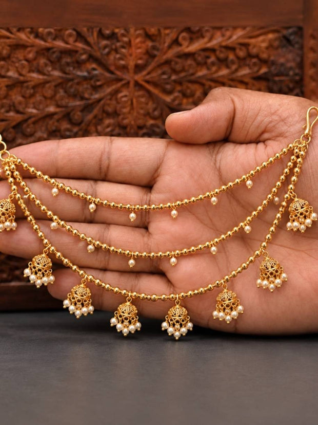 Gold-plated three-strand jhumka ear chain with pearl drops and filigree charms held in a woman's open palm against carved wooden backdrop