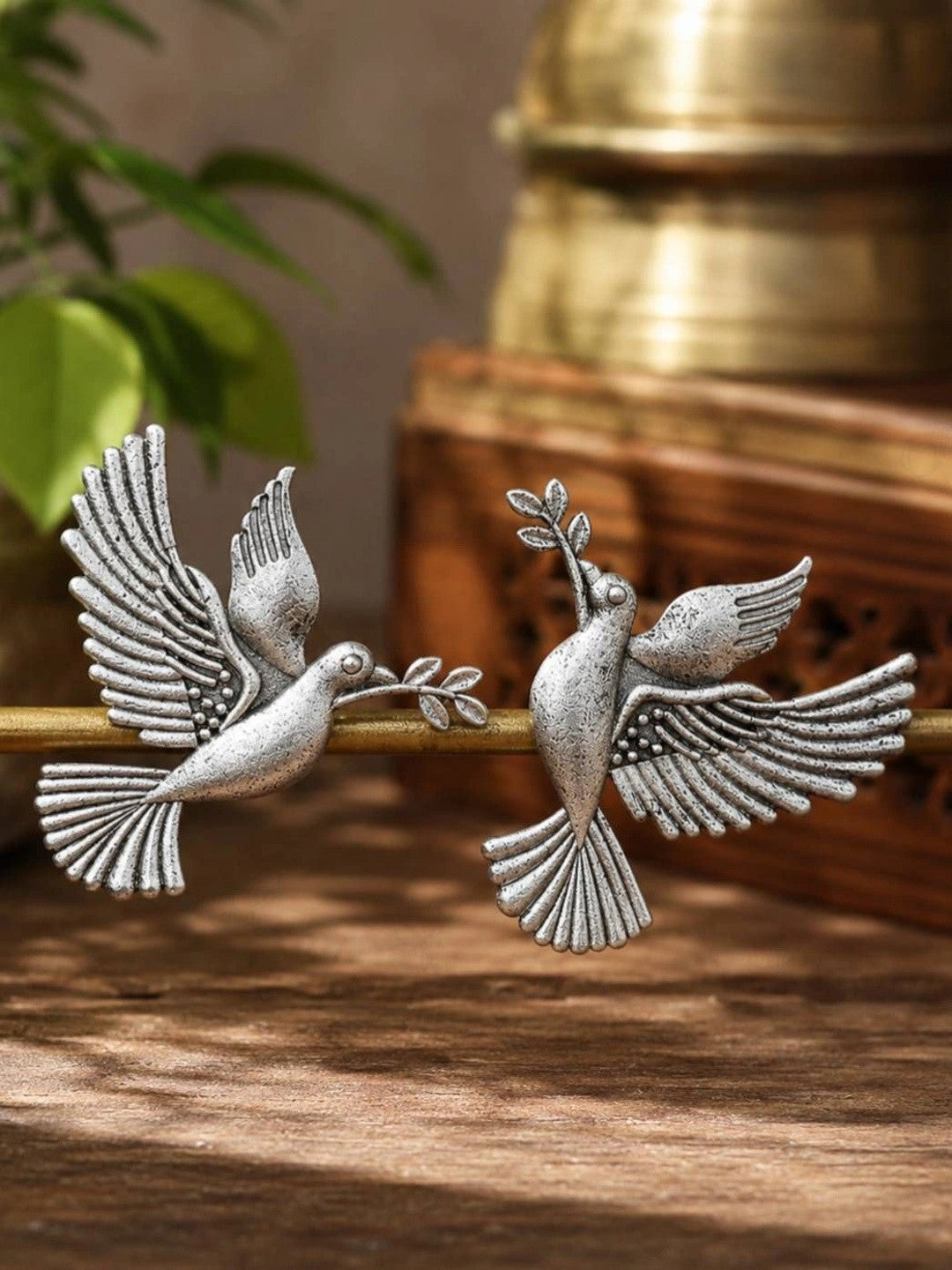 Pair of oxidised silver dove earrings displayed on a brass rod with wooden and plant backdrop